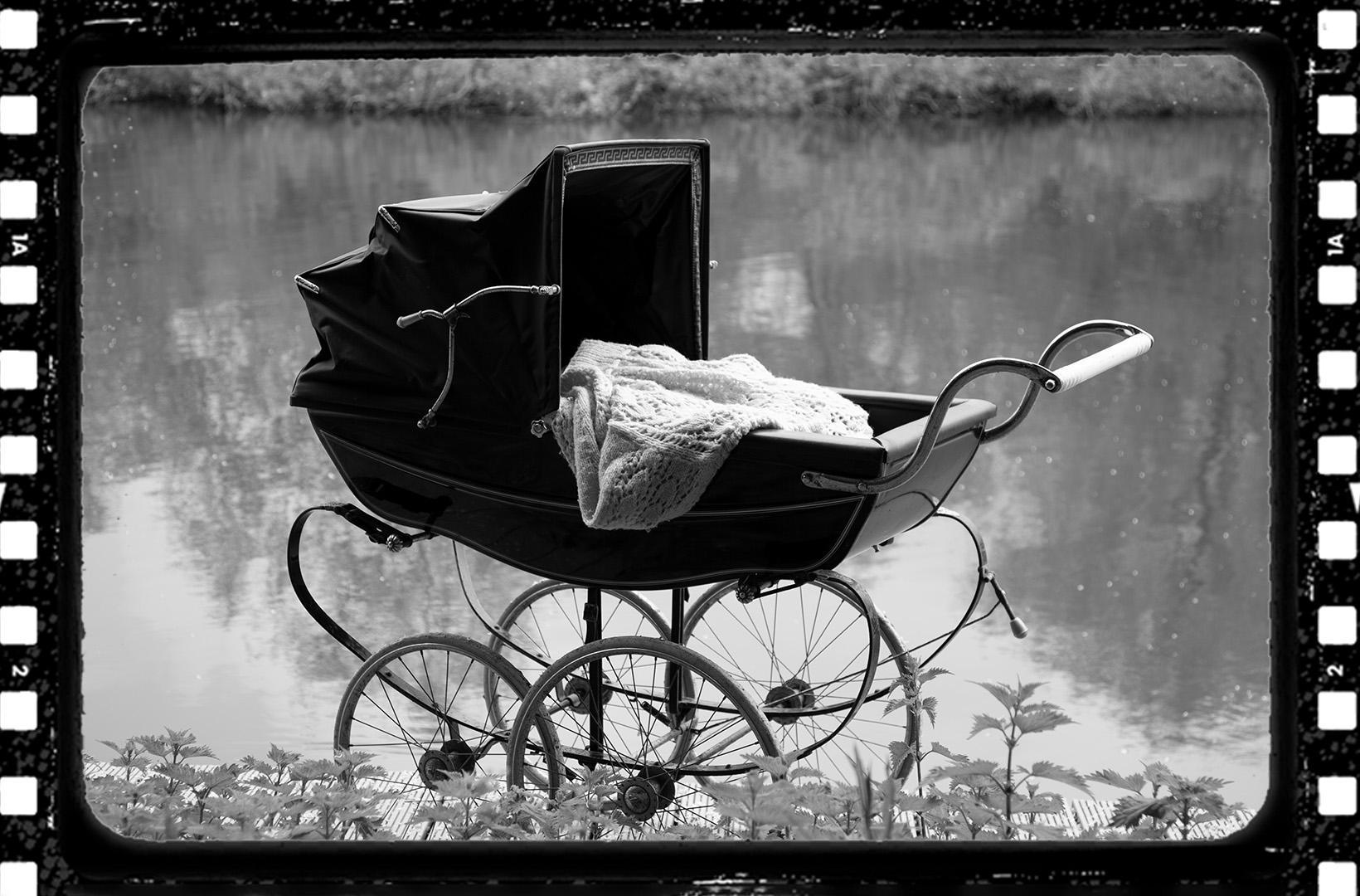 Black and white photo of an old fashioned babies' pram, empty, next to a river. A celluloid film frame has bee overlaid onto the image to make it look cinematic. 