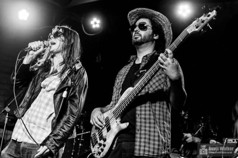 Black and white photo of two musicians standing facing forward and slightly to the left. Both wear shirts over t-shirts and sunglasses. One is singing into a mic and the other is playing guitar .