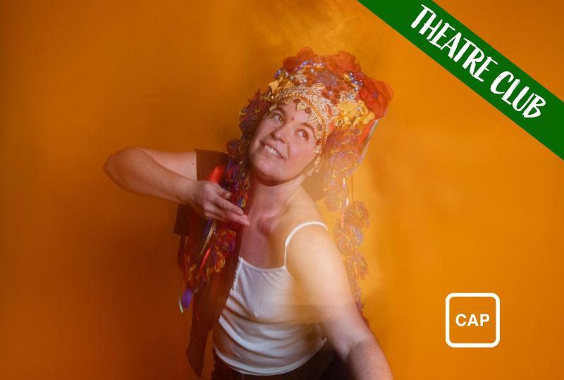 Photograph of performer Katherina Radeva, wearing a white vest top and elaborate gold and red headdress decorated with jewelled flowers. She is mid-dance, holding one hand up to her face and looking upwards. She is standing in front of an orange backdrop, and there is a blurred camera effect across the image, giving the impression of movement and reflection. White text reading 'Theatre Club' is overlaid across the top right corner and the symbol for Captioned Performance is in the bottom right corner
