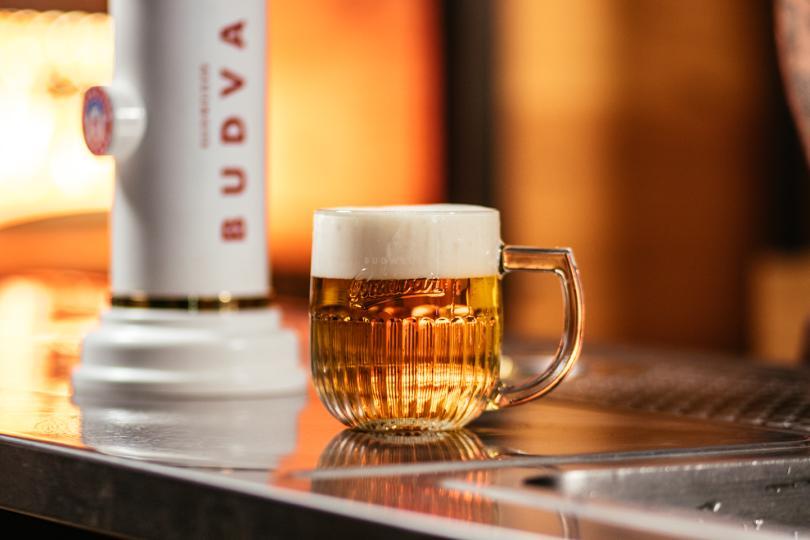 A pint of golden lager with a foamy head sits in a handled glass on a shiny bar
