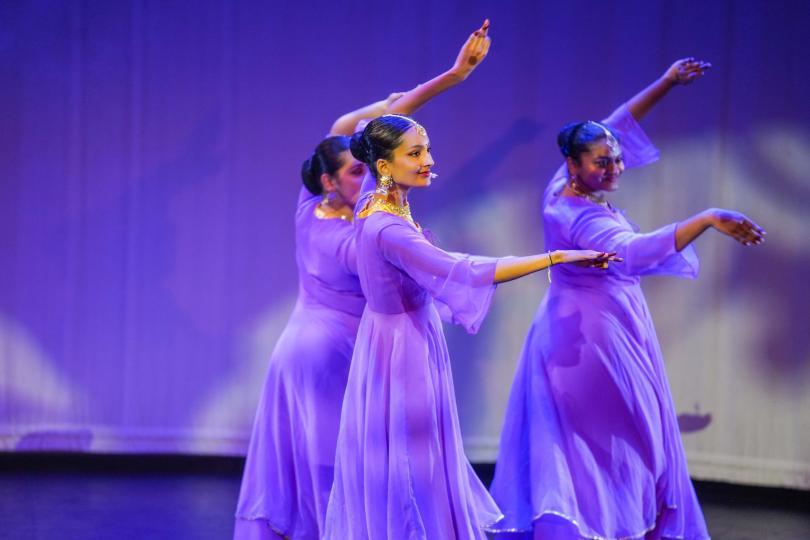 Three South Asian women in purple dresses performing classical dance poses