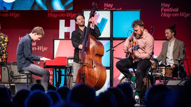 four musicians on stage. Left to right, they play electric piano, upright bass, electric guitar and drums.