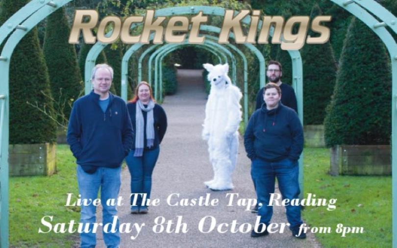 Five figures stand on a path facing the camera. The path stretches straight back into the distance in the centre of the image; it has a series of teal arches over it and sculpted evergreen trees on grass either side. Three of the figures are wearing blue jeans and navy blue comfy jumpers, the head of a fourth figure is visible almost directly behind one of the front individuals and finally at the back of the group a fifth figure is dressed head-to-toe in a white furry animal suit