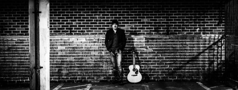 Photo of Craig Joiner with his guitar. Craig is standing in front of a wall in an urban street. The sky is dark and Craig is lit up by a street lamp. The photo is in black and white, giving it a retro feel.