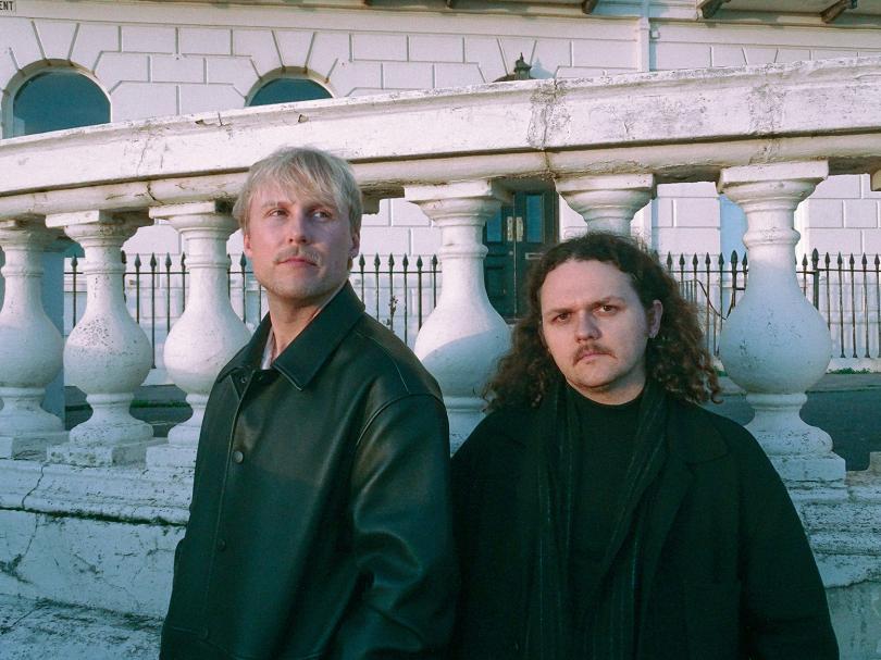 Puma Theory band members standing outside a stately home. One has short blonde hair and a blonde moustache, the other long brown hair and a dark moustache.