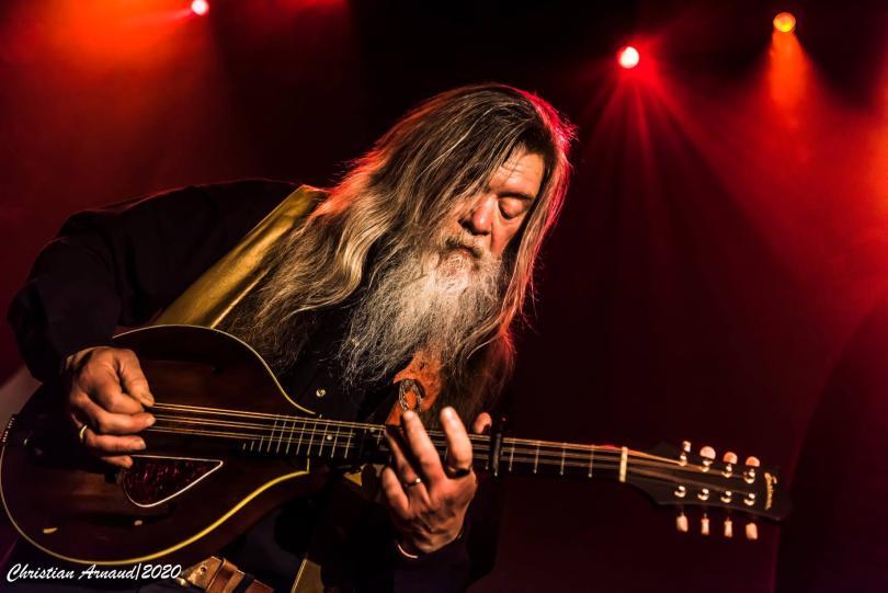 Person playing a mandolin. The background is dark but with patches illuminated by red stage lights. The musician is dressed in dark clothing and has long straight hair and a long white beard