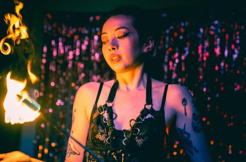 A tattooed, femme presenting performer poses in front of a shining purple glitter curtain. They wear a sultry black lace garment, and are holding flaming swords as part of a fire eating act