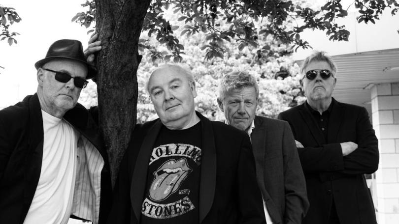 Black and white line up of 4 musicians all looking towards the camera and wearing dark jackets. The individual on the far left has dark glasses and a hat and leans on the trunk of a tree which shelters the whole band. The next person is bald and wears a Rolling Stones T-shirt, the third member of the band is slightly behind him and then the final member also wears dark glasses and has his arms crossed.