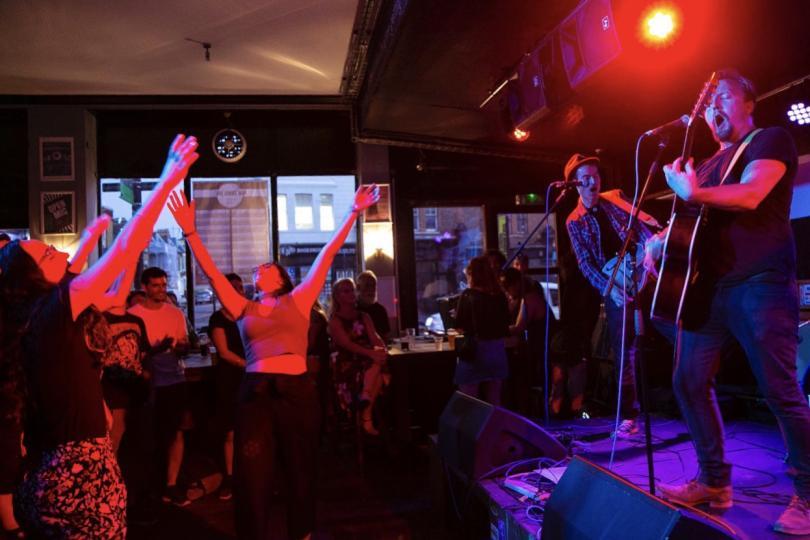 A pub gig; two musicians are on a stage on the right of the image, they are both wearing jeans, playing guitars and singing and bathed in violet light. Centre and left of the image shows the audience who are bathed in red light and have their arms in the air.
