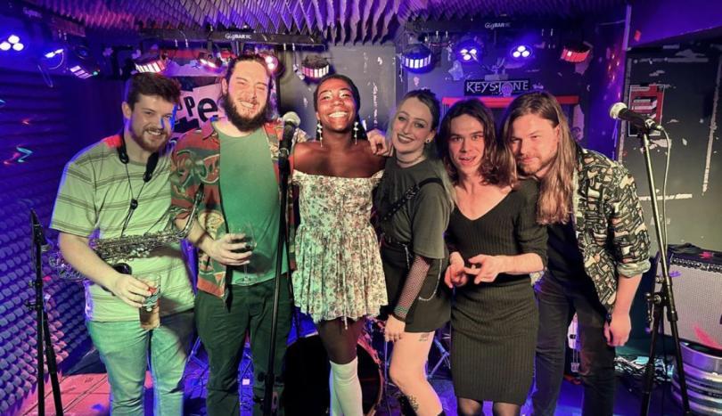 Six musicians with big smiles are standing leaning in towards each other facing the camera. They have blue lights overhead, three microphones in front and an amp to one side.
