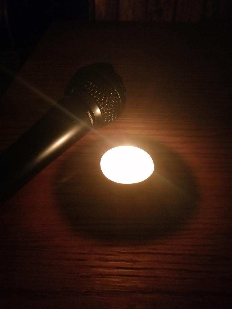A tealight candle shines in a dark space illuminating a wooden surface; just on the edge of the pool of light is a black microphone