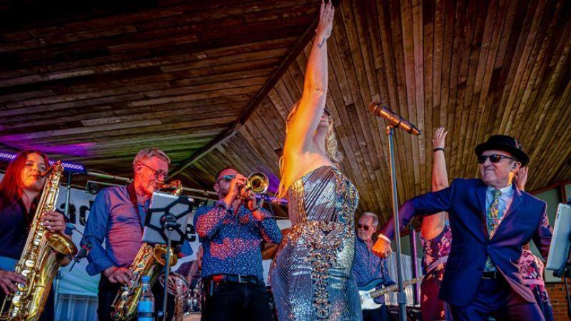 The image shows Madison Heights on the stage performing. The singer is dressed in a silver dress and the brass section of the band can be seen behind her.