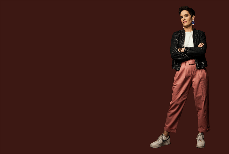Photograph of Jen Brister, wearing a white t-shirt, black sparkly jacket, dusky pink trousers and white trainers. She is standing with her arms crossed against a dark red backdrop.