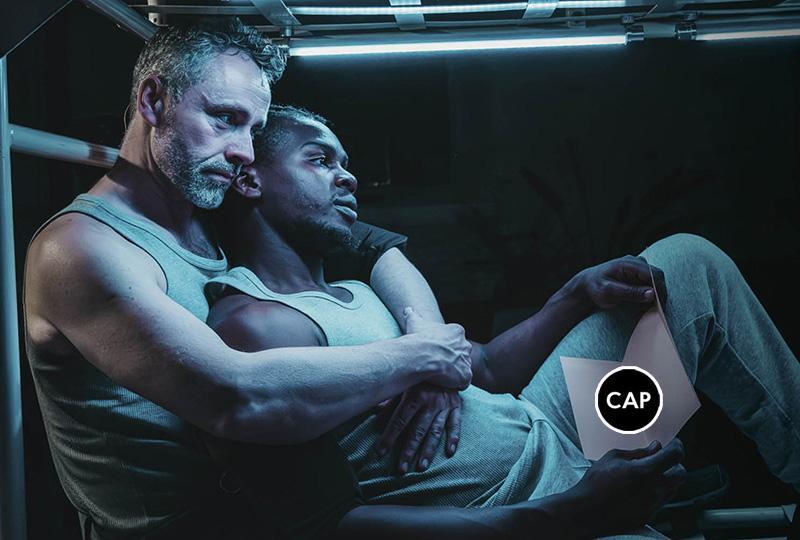 Photo from a performance of Kiss Marry Kill. Two prison inmates wearing grey sweatpants and tank tops are sitting on the bottom bunk of a prison bed cuddling. One is holding a letter. The symbol for 'Captioned Performance' is in the bottom right corner of the image.