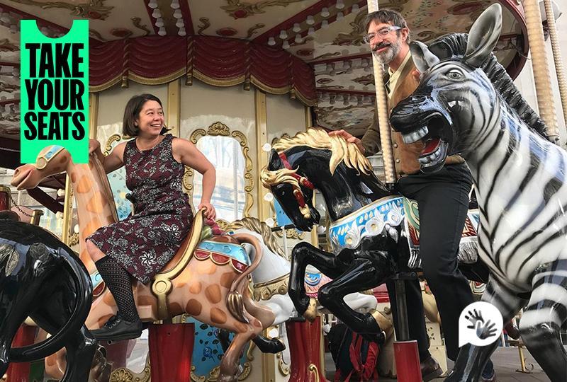 Photograph of performers Jo Fong and George Orange riding an old fashioned carousel, sitting on models of a giraffe and horse respectively. 'Take Your Seats' logo is in the top left corner and the British Sign Language symbol is in the bottom right corner.