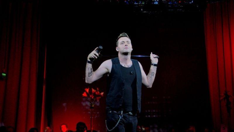 The image shows a Robbie William tribute artist stood in front of a red curtain.