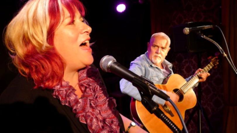 Sally Ironmonger and Brian Carter performing. Sally is singing and has a smile on her face. Brian is playing a guitar and looking towards Sally.