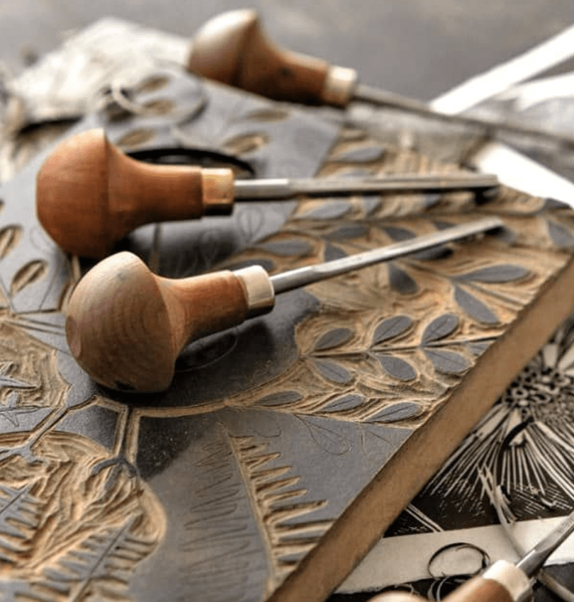An image of a carved lino block with ink on. On top of the block are two carving tools with wooden handles.