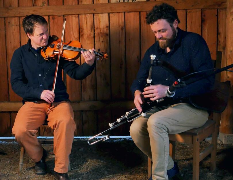 Photo of Bradon and Dominic, the two members of Straight from the Jug sitting on a bench with a wood-clad wall behind and playing fiddle and uileann pipes.