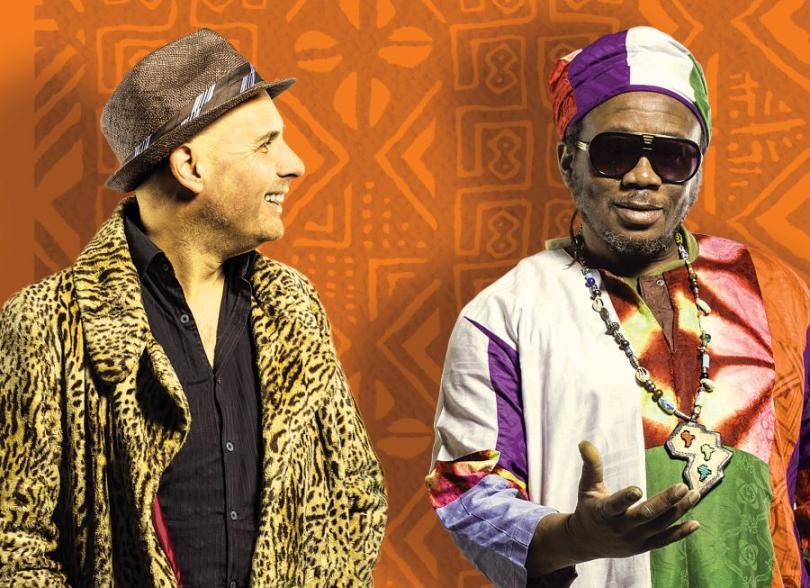 A patterned orange backdrop with Two musicians shown from the waist up.  The figure on the right looks towards us, he is black with a little bit of facial hair and is wearing large sunglasses and multi-coloured clothing. The figure on the left has turned his head to face his companion, he is white with stubble and wears a leopard print jacket.