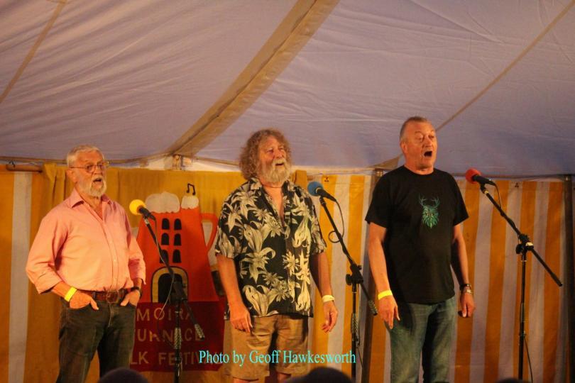 Image of the three members of The Laners singing in a folk club setting. The trio are dressed informally.