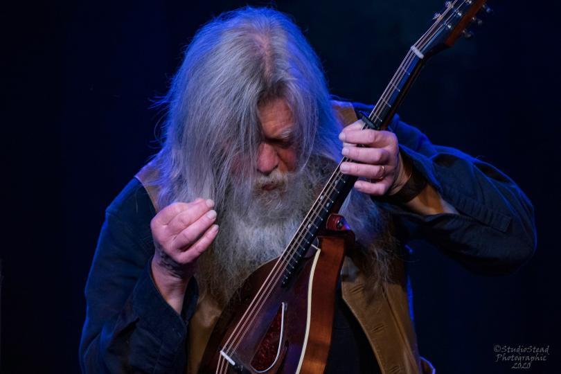 Grey haired and bearded man intently focuses on playing his mandolin against a dark blue background