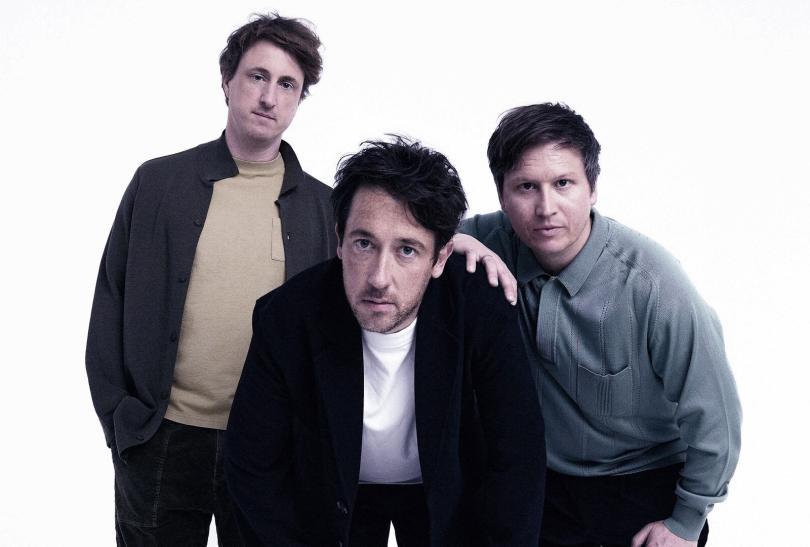Band The Wombats, three white men in their late thirties wearing light jackets and t-shirts, looking puzzled at the camera