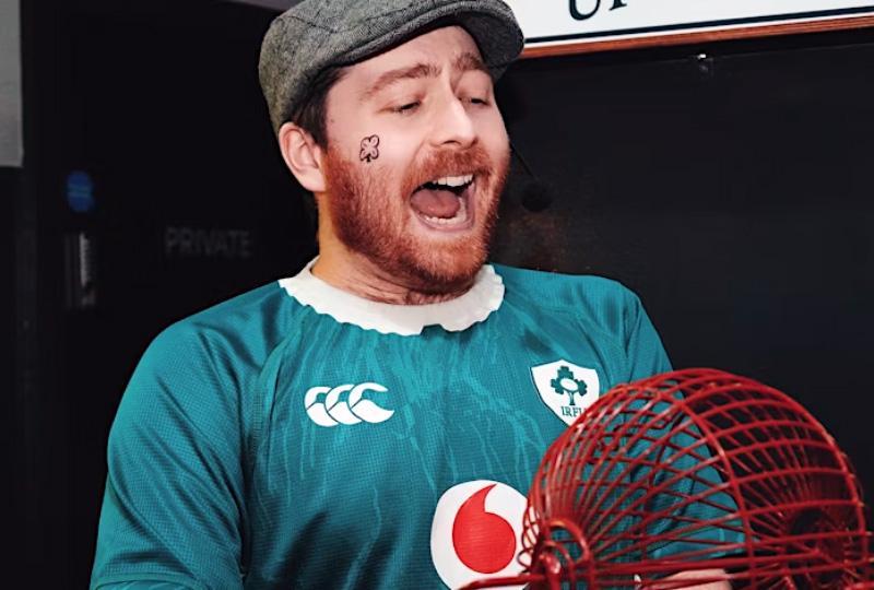 Man with a ginger beard and a big grin, wearing a flat cap and Ireland football shirt, holding a bingo wheel