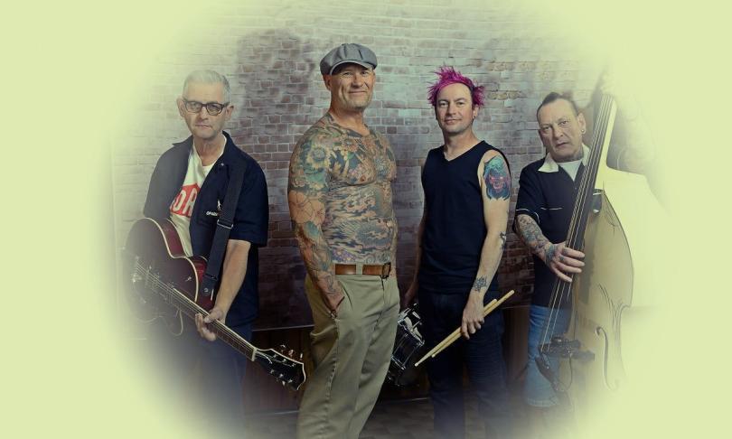 Photo of the band members of Guana Batz, four middle-aged men. Two are holding instruments, one is shirtless and covered in tattoos.