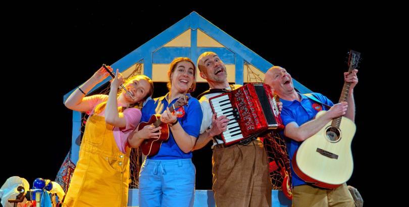 Four actor-musicians on stage wearing beach-style costumes in front of a beach hut