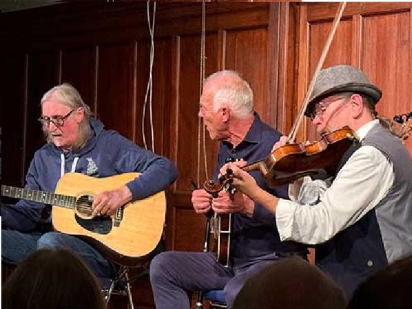 Photo of the three musicians in Baum, Maggs and Taylor. They are sitting together on stage and playing guitar, mandolin and fiddle.