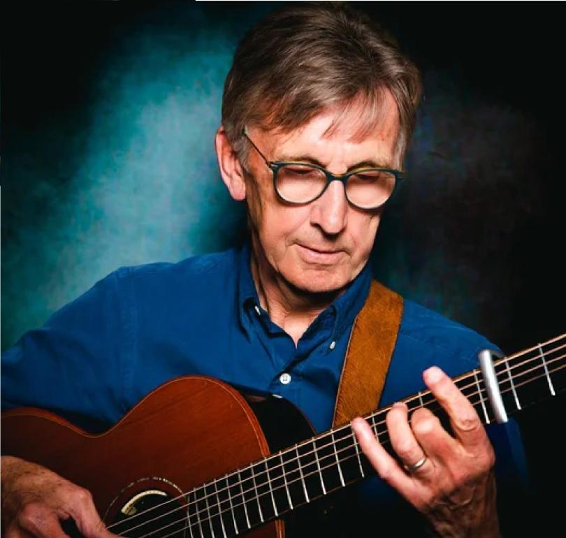 Photo of Bob Wood playing guitar. There is a spotlight on Bob's face and hands. The background is lit in blue and Bob is wearing a blue shirt.