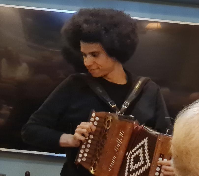 Photo shows Cohen Braithwaithe Kilcoyne playing at a folk club. Cohen is playing a melodeon. He is looking down to the side and smiling.
