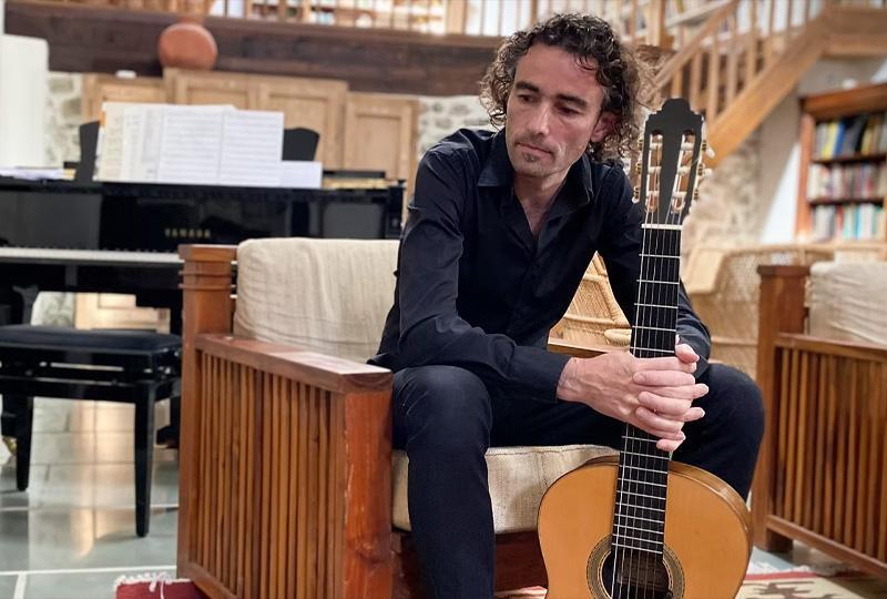 Guitarist Gerard Cousins sat holding his acoustic guitar in a library-like room with a piano in the background