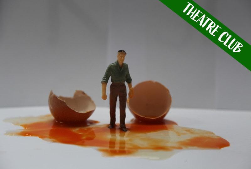 Photograph of a miniature figure of a man wearing a dark shirt and trousers, stood on a white table surface in the centre of a pool of egg white and yolk. There are two halves of a broken eggshell on either side of the figure. White text reading 'Theatre Club' is overlaid on the top right corner