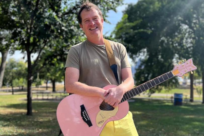 Singer Dick Valentine in a grey t-shirt and yellow shorts, smiling and holding a pink guitar