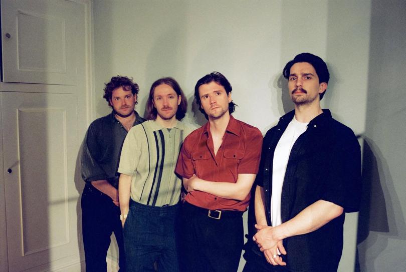 Members of the band "Do Nothing", four twenty-something men with hipster haircuts and moustaches, standing in front of a door