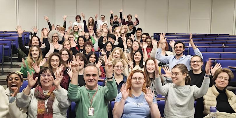 Auditorium filled with people of various ages all waving their hands at the camera