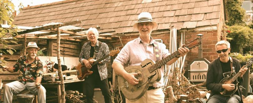 Four older white male musicians  outside a brick and slate building with firewood around. 1st and 3rd musicians in the row wear hats. Left to Right they play: Percussion, bass, National steel guitar, mandolin