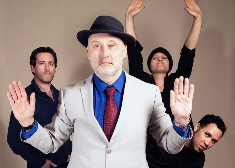 Band members standing with their hands up. Singer Jah Wobble is at the front in a white suit jacket and trilby hat