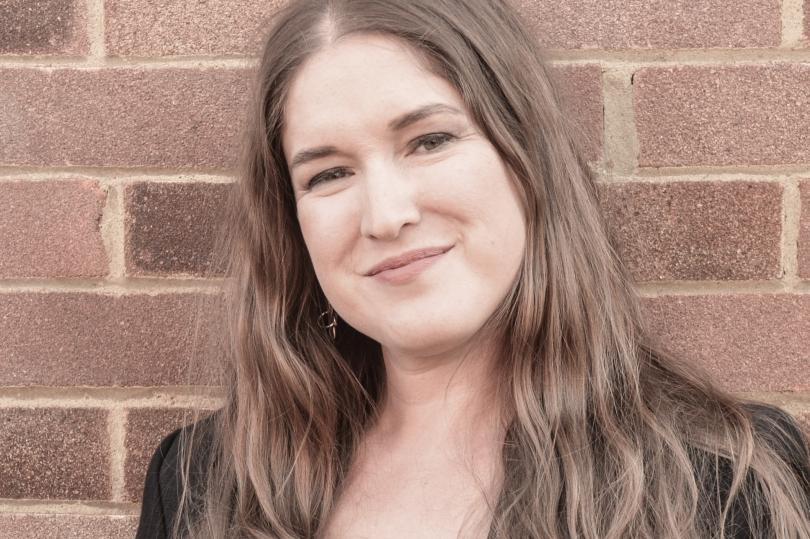 Singer Jessica Lee Morgan with long brown hair and a big smile, standing in front of a brick wall