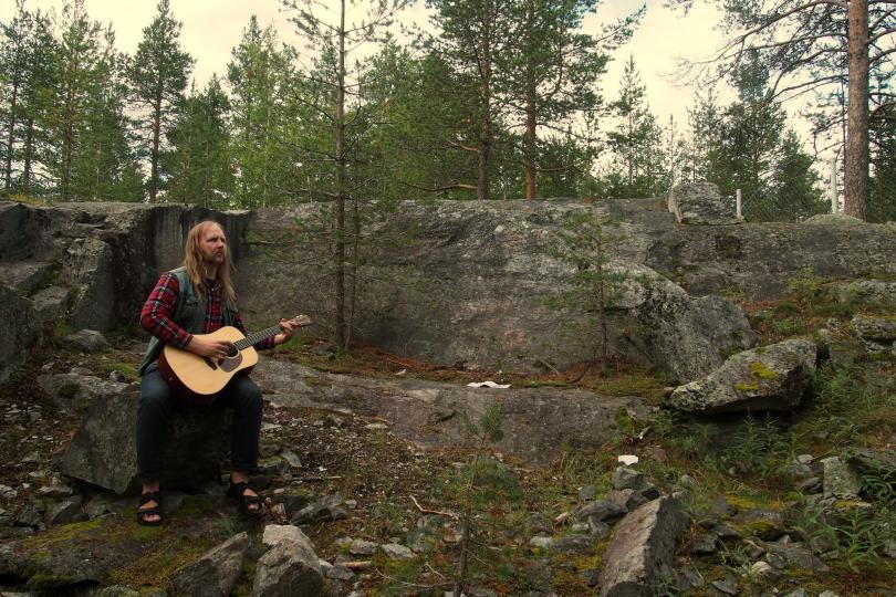 Long-haired musician wearing chequed flannel shirt, denim waistcoat, jeans and sandals with an acoustic guitar sits on a rock to the left of the image in front of a stony outcrop in pine wood.