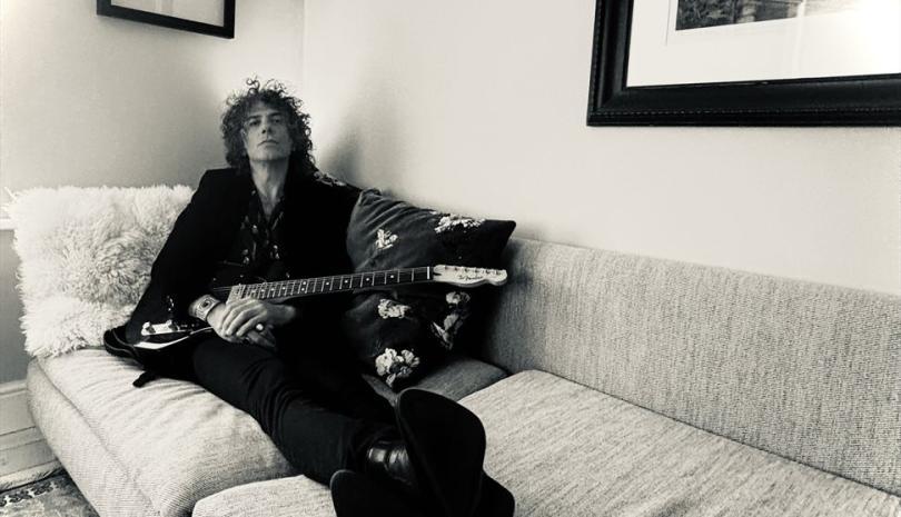 Kavus Torabi in a black suit, lounging on a white sofa with a guitar
