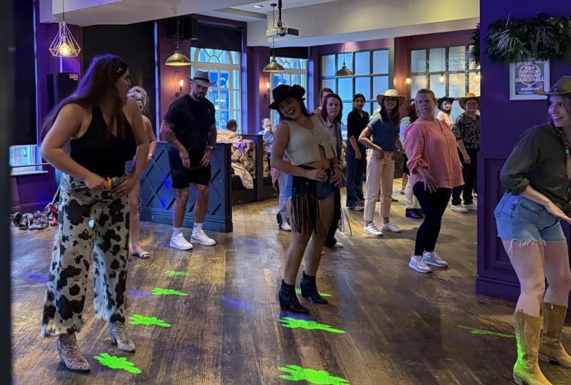 Group of people learning line-dancing steps in a bar. They are mostly wearing cowboy hats.