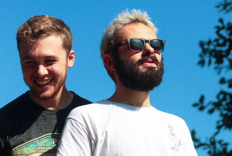 Two musicians are shown from the waist up with a strikingly blue sky behind them. The one on the left of the image wears a grey t-shirt, they have pale skin, short ash-brown hair, short thinnish facial hair and their eyes crinkle in a smile. The person on the right of the image is wearing a white t-shirt and sunglasses; they also have pale skin, their hair is short bleach blonde and they have a short dark beard