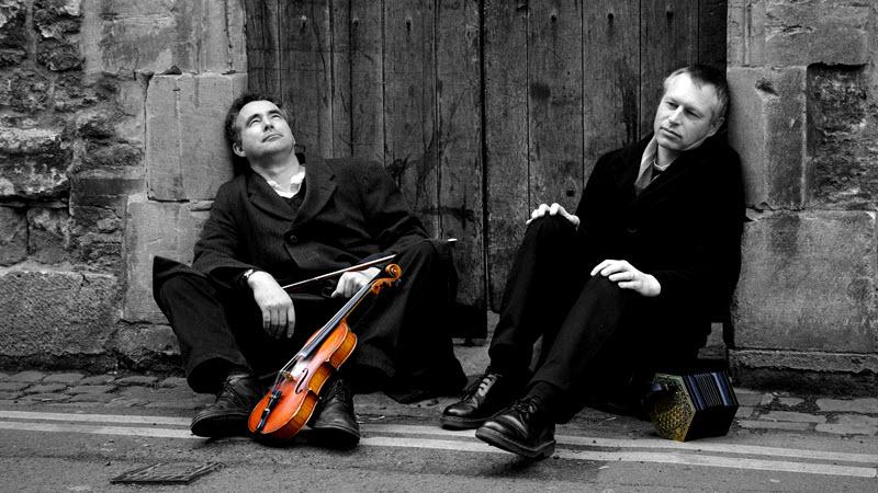 Black and white photograph of Mat Green and Andy Turner sitting in a nonchalant pose on the ground in front of an old wooden door and over double-yellow street lines. Mat has a fiddle, the only coloured object in the photo, and Andy has a concertina.