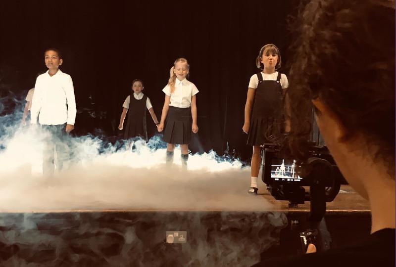 Children on stage with dry ice, being filmed by a digital camera