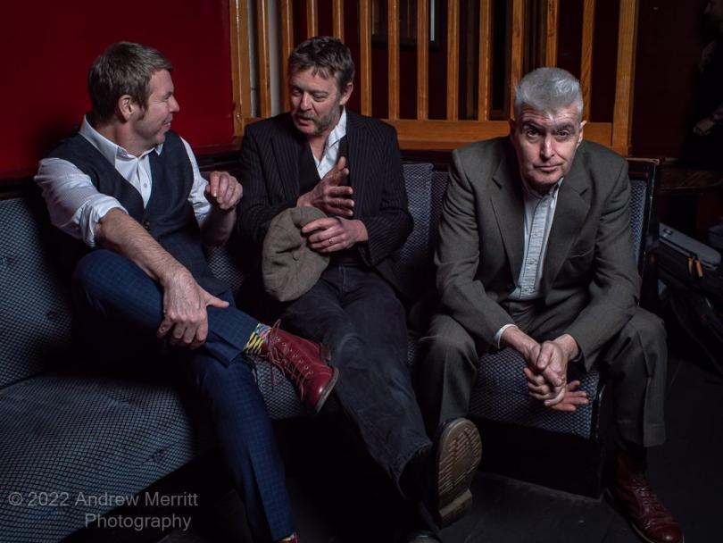 Three bandmates sit on a corner sofa. All are dressed smart casual with open neck shirts and a hint of rockabilly. The two on the left are chatting to each other and the one on the right is staring at the camera quizzically