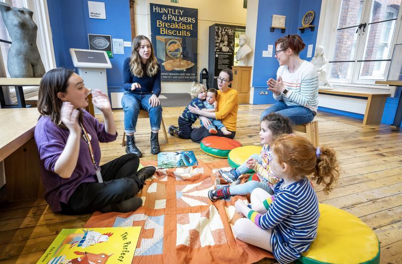 Rhymetime at Reading Museum, member if staff is sat on a patchwork quilt surrounded by toddlers and their parents
