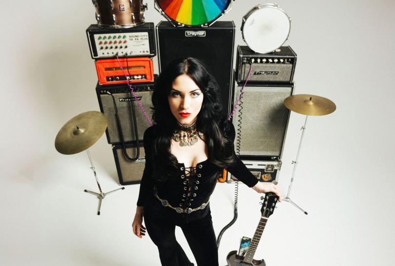 Rosalie Cunningham wearing a black velvet jupsuit standing in front of a stack of amps and instruments